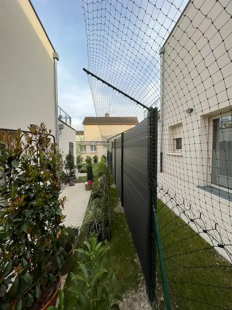 Enclos extérieur en grillage métallique vert avec toit en grillage, entouré de haies, dans un jardin avec pelouse sous un ciel bleu.