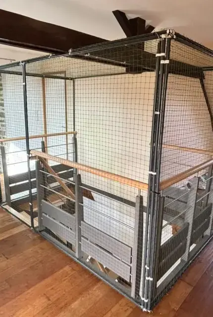 Structure grillagée encadrant une montée d'escalier intérieure d'une maison avec un sol en parquet.