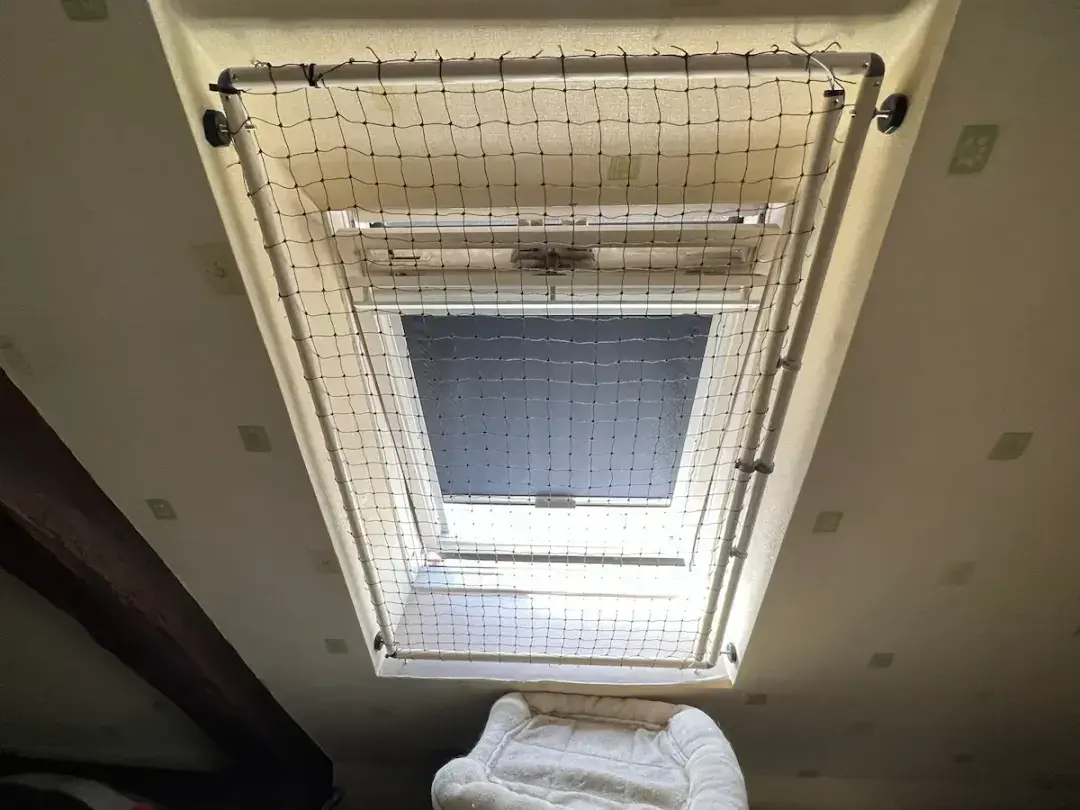 Une fenêtre de toit ouverte avec une moustiquaire sur un plafond incliné dans une pièce minimale avec une table et des vêtements pliés.